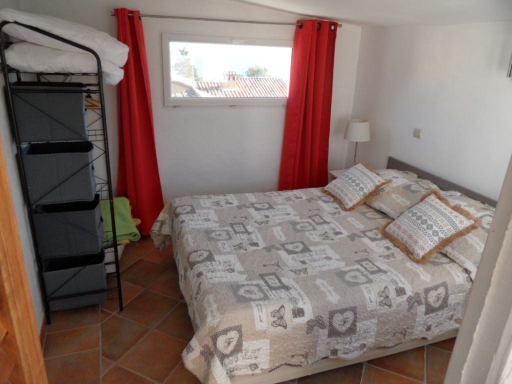 Een slaapkamer met een bed, een raam met rode gordijnen, een bedlampje en een klein rekje.