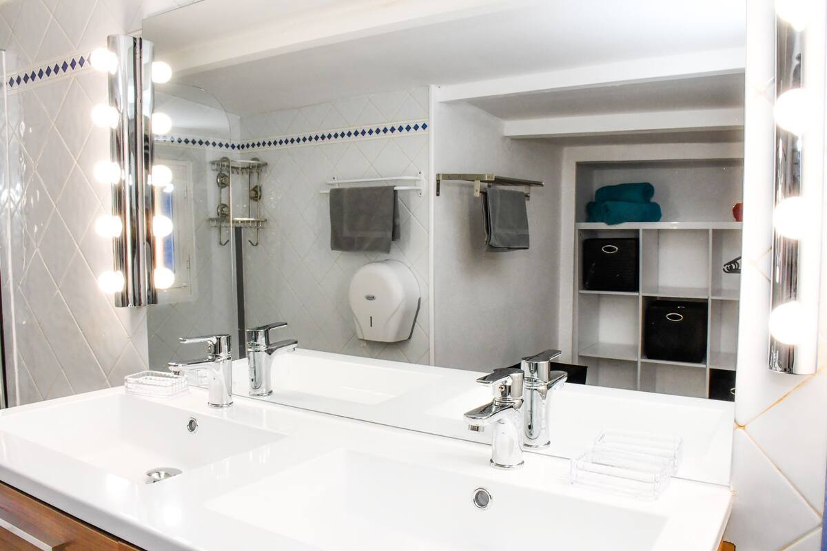 Une salle de bain moderne avec deux vasques, un grand miroir avec éclairage intégré, un porte-serviettes et une étagère contenant des articles de toilette.