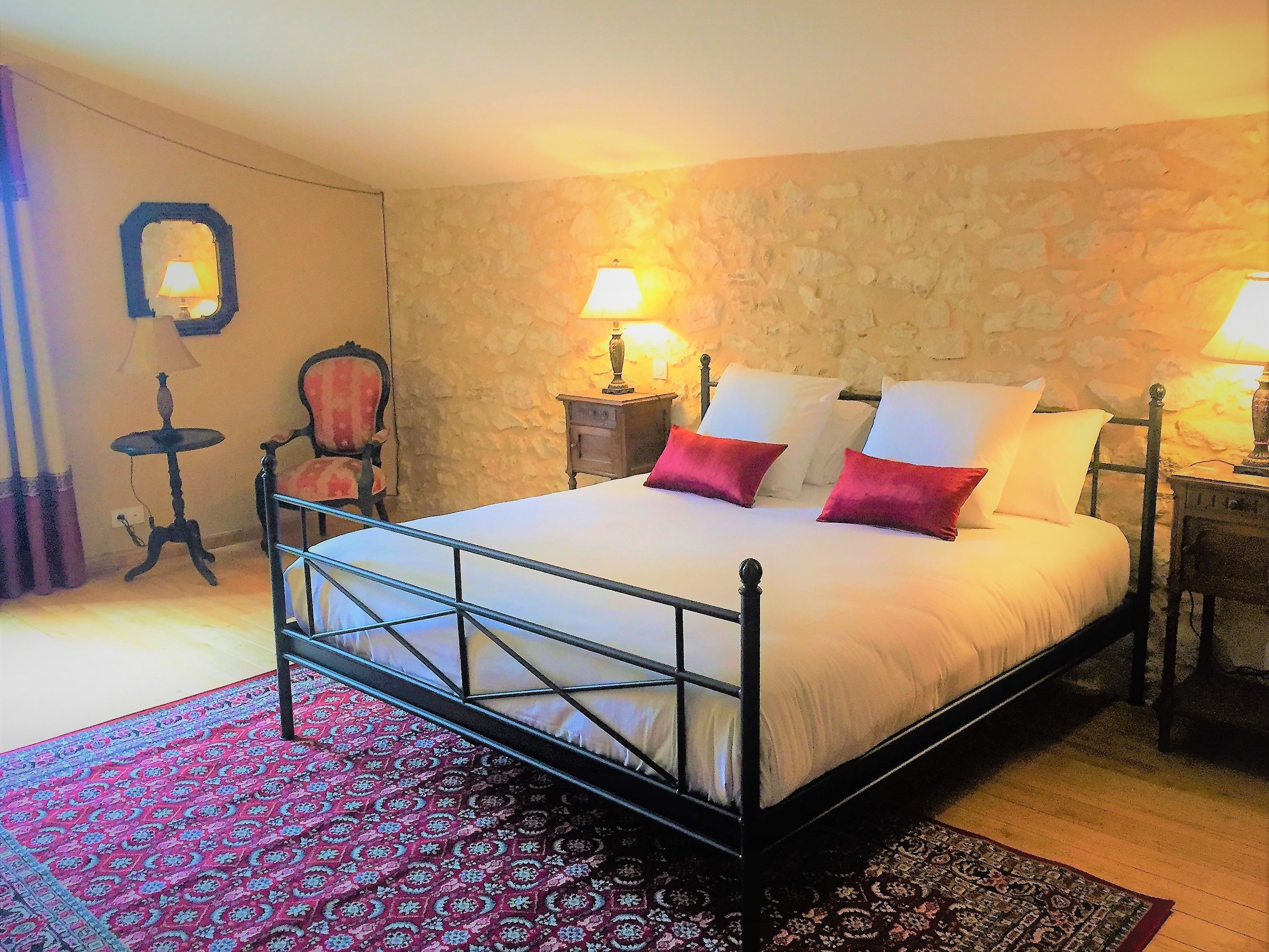 Un dormitorio con pared de piedra, cama con ropa blanca y almohadas rojas, dos lámparas de noche, una silla y una alfombra estampada.