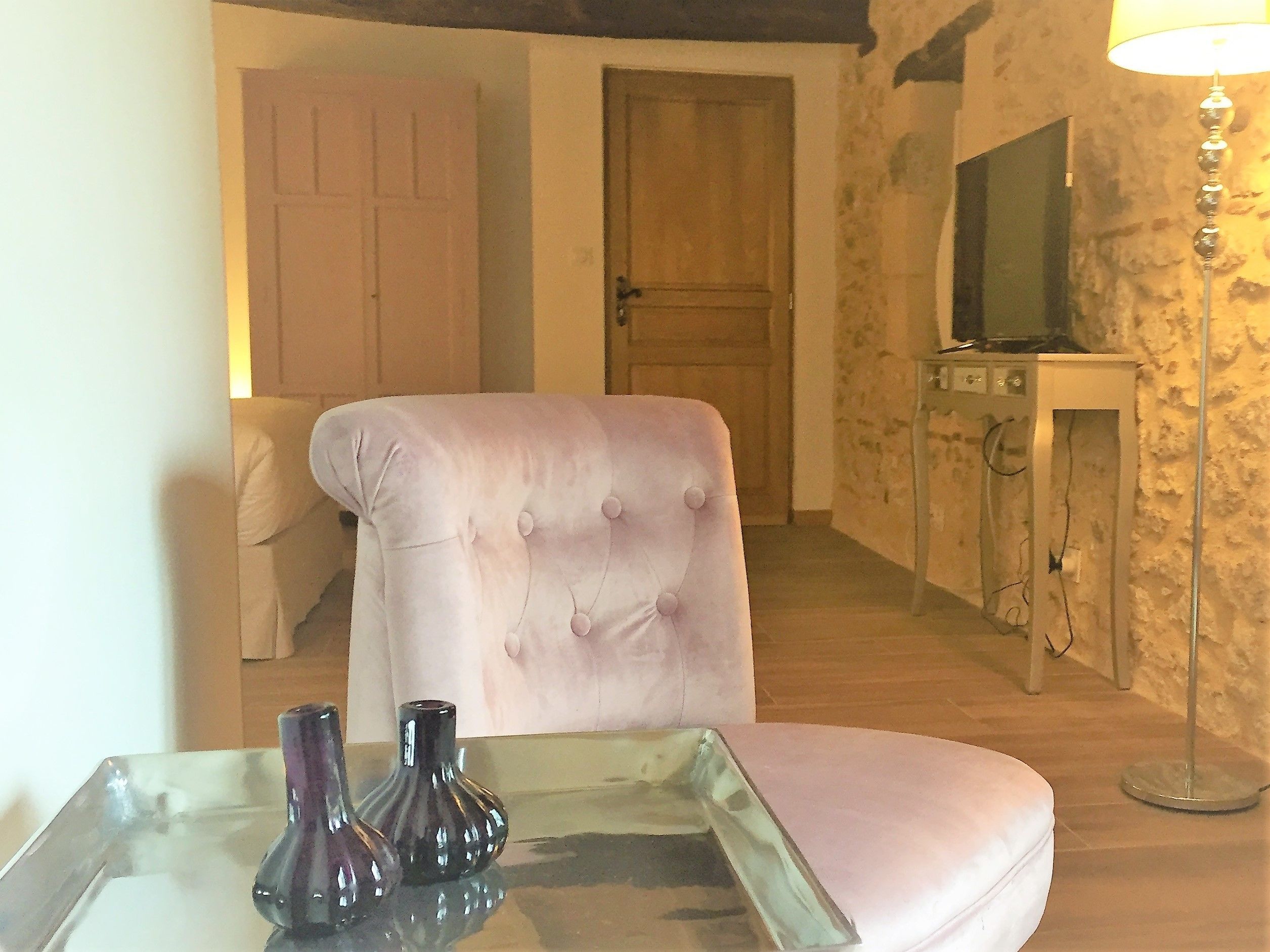 A room with a tufted armchair, a small table with decorative vases, a wooden door, a stone wall, and a floor lamp.