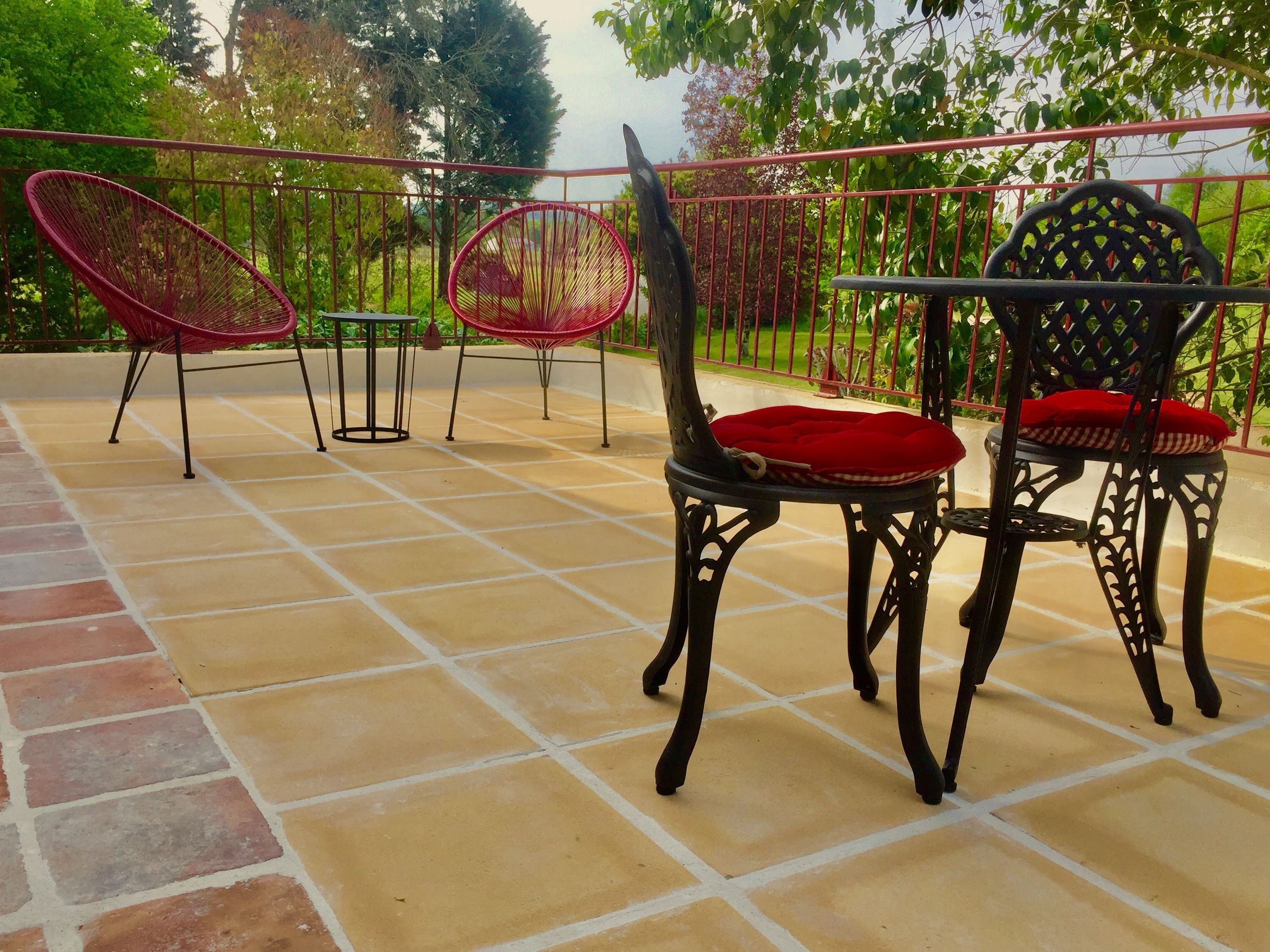 Une terrasse avec un sol carrelé et une rambarde en métal. On y trouve trois chaises en osier avec des coussins rouges et une chaise en métal noir avec un coussin rouge.