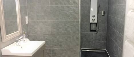 Baño con lavamanos blanco, espejo y toallero. Se observa una ducha con panel de control y una cesta blanca.