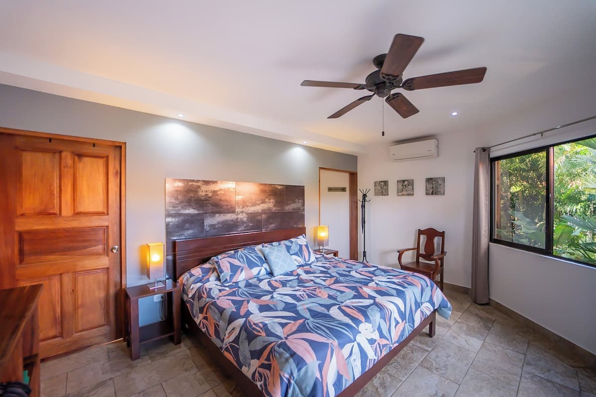 A bedroom with a bed, a ceiling fan, a wooden door, a nightstand with a lamp, and a window with a view of greenery.
