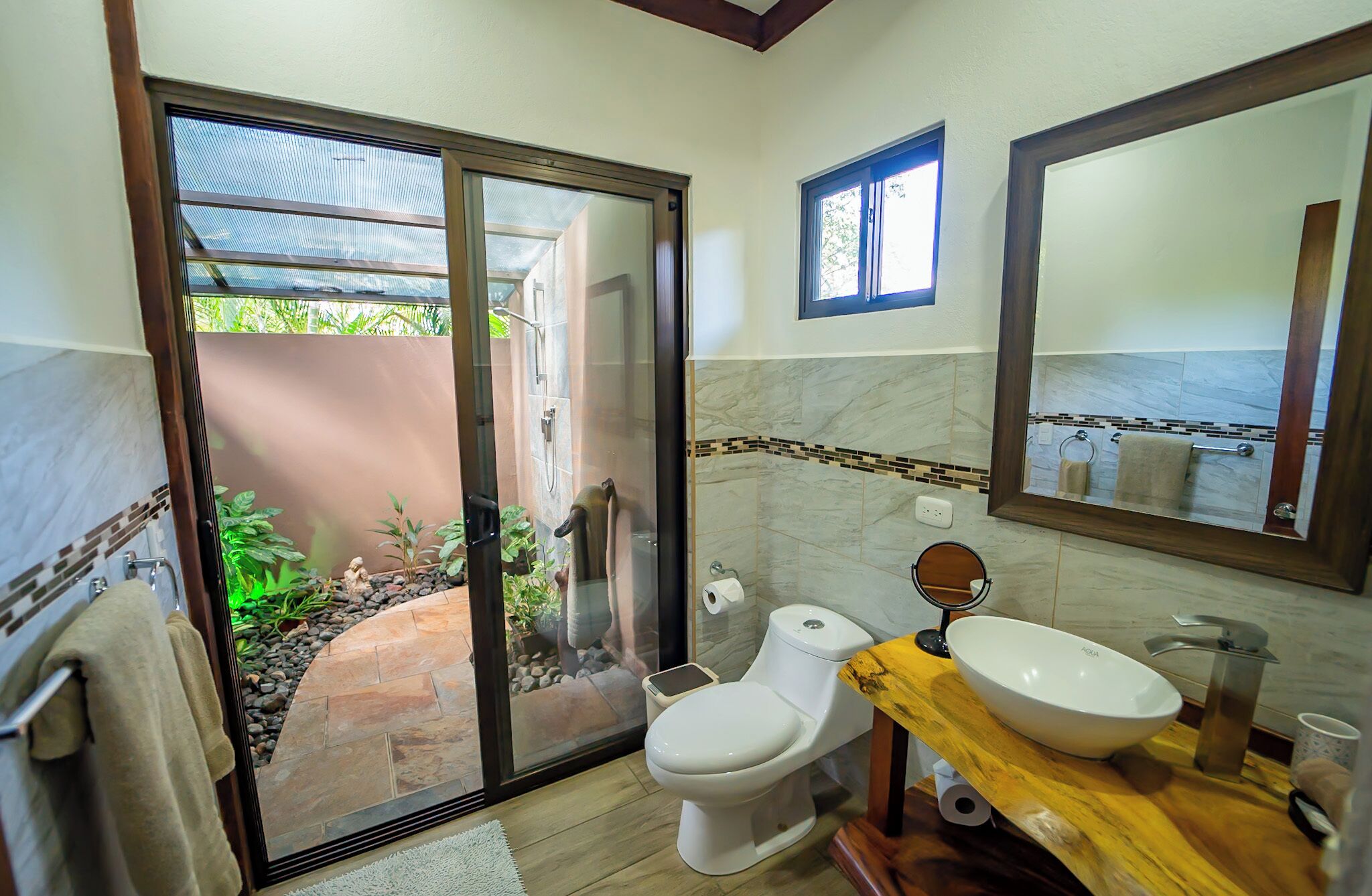 A bathroom with a toilet, sink, and large mirror. There is a sliding glass door leading to an outdoor area with plants and a statue.