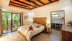 Un dormitorio con una cama grande, vigas de madera en el techo, un ventilador de techo, un gran ventanal corredero que da a un balcón con vegetación, una pequeña cocina y suelo de madera.