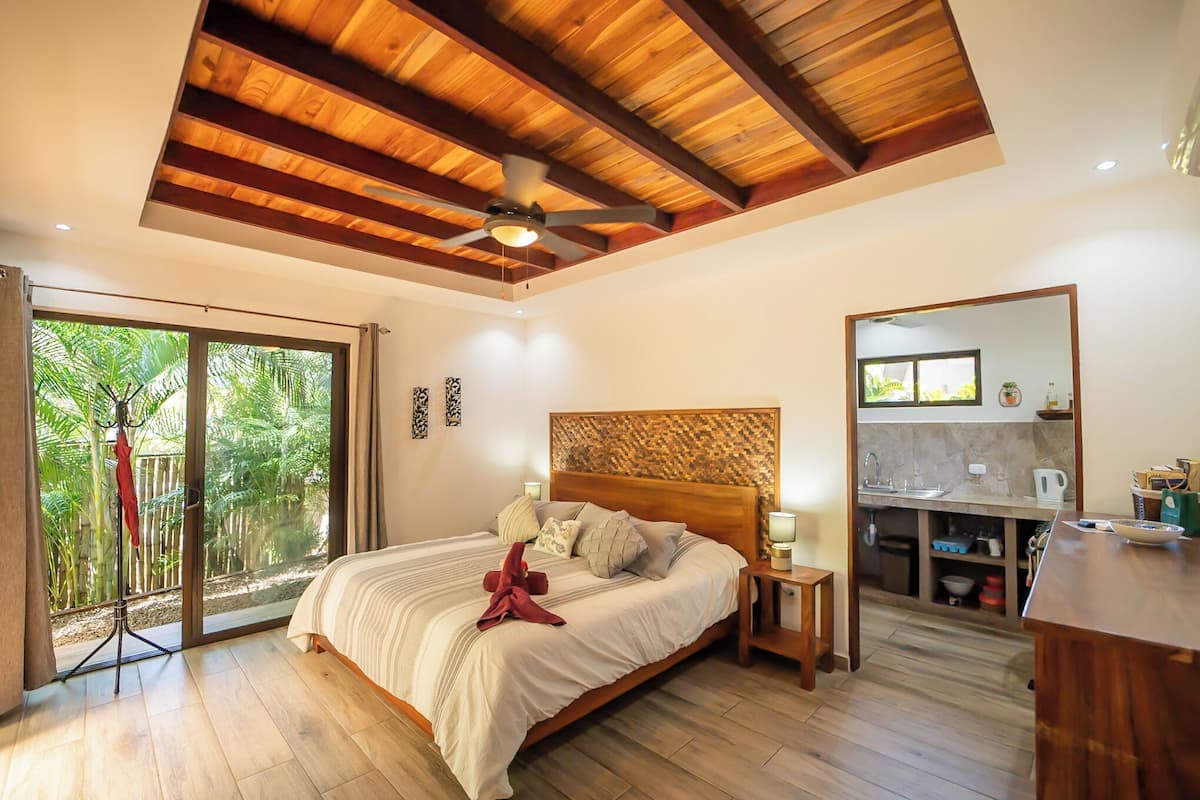 A bedroom with a large bed, wooden ceiling beams, a ceiling fan, a sliding glass door leading to a balcony with greenery, a small kitchen area, and a wooden floor.