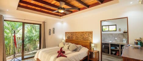 A bedroom with a large bed, wooden ceiling beams, a ceiling fan, a sliding glass door leading to a balcony with greenery, a small kitchen area, and a wooden floor.