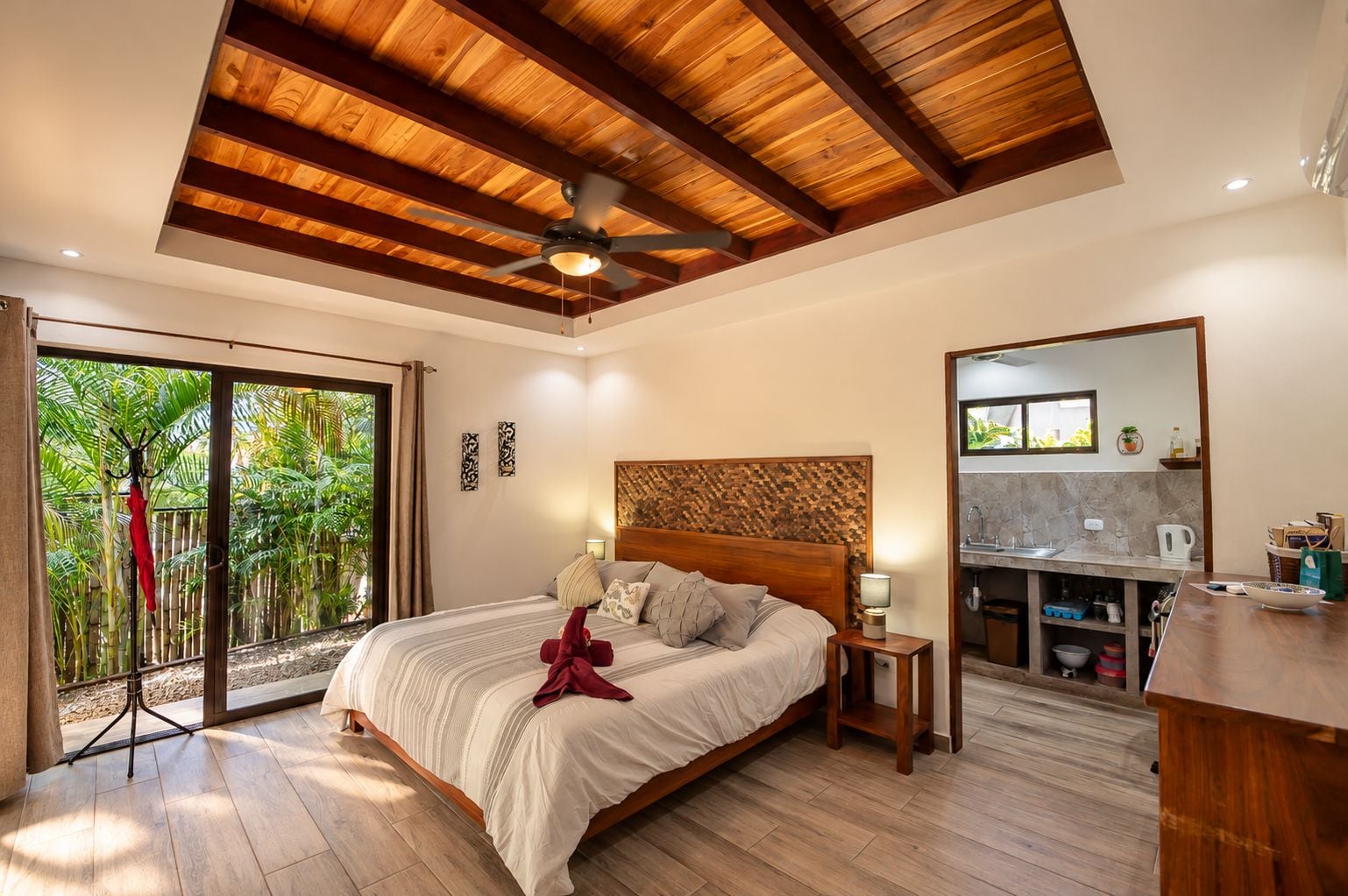 A hotel room with a bed, a ceiling fan, a view of greenery through a sliding door, and a kitchenette.