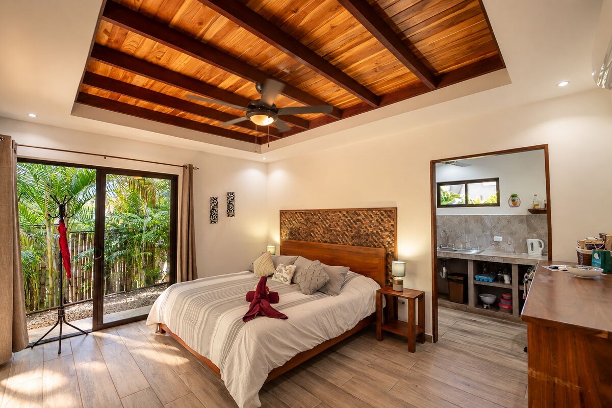Habitación de hotel con cama, ventilador de techo, vista a vegetación a través de una puerta corrediza y una kitchenette.