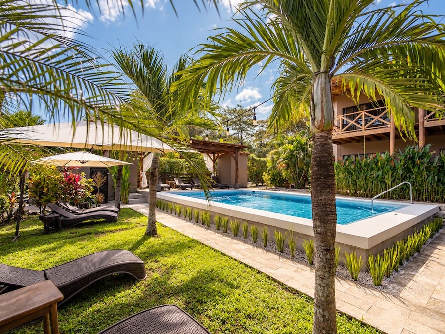 Un área de piscina con sillones de descanso, palmeras y una zona de estar cubierta.