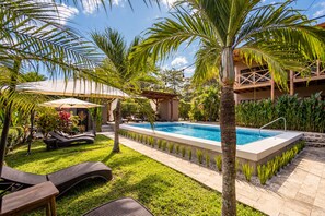 Un área de piscina con sillones de descanso, palmeras y una zona de estar cubierta.