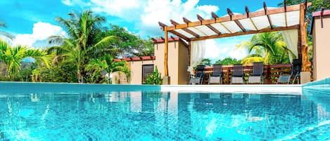 A clear swimming pool with a wooden pergola and lounge chairs, surrounded by palm trees and a clear sky.
