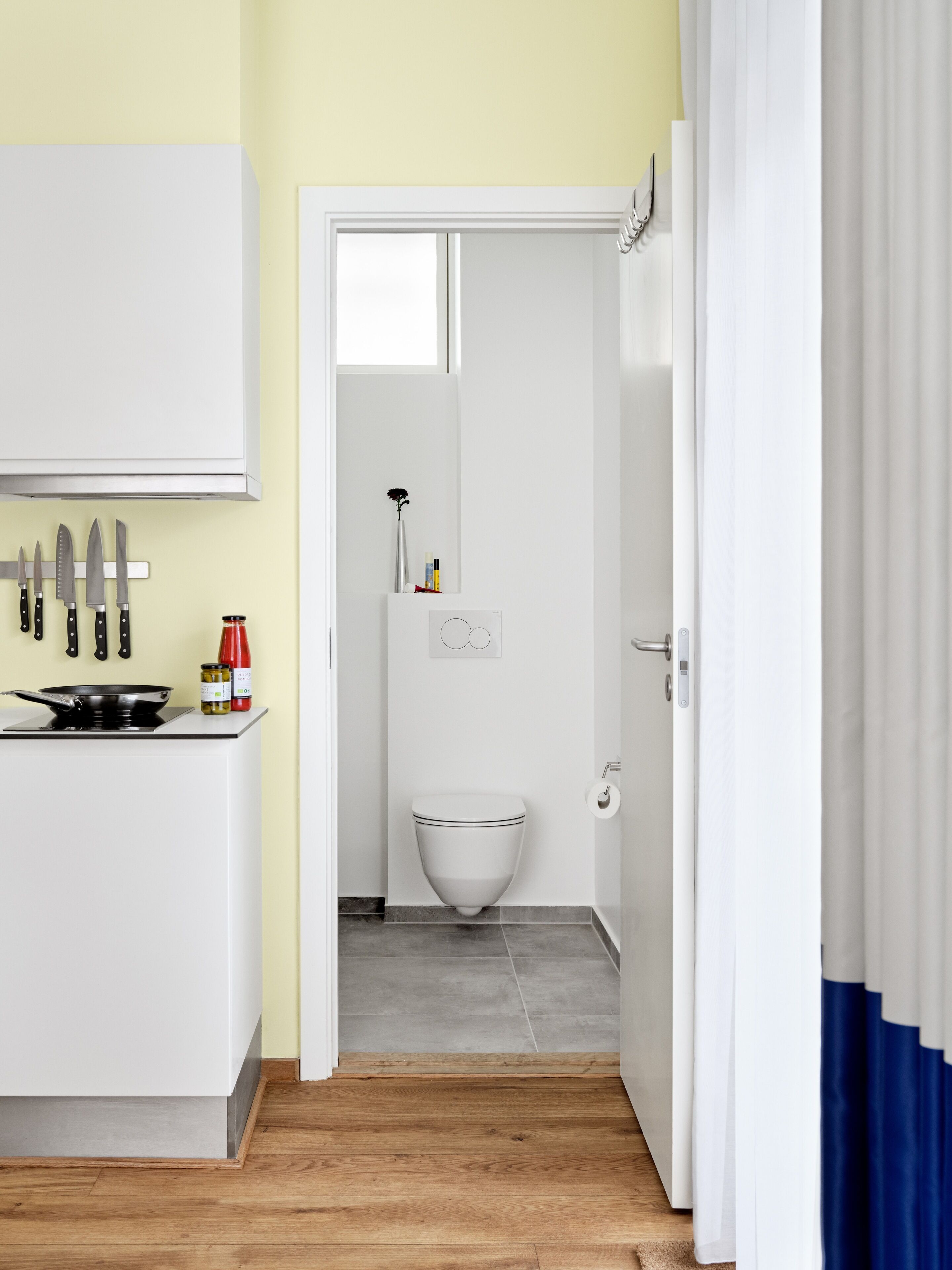 A compact kitchen with a stove, a sink, and a countertop with knives and condiments. A white toilet is visible in the adjacent bathroom.