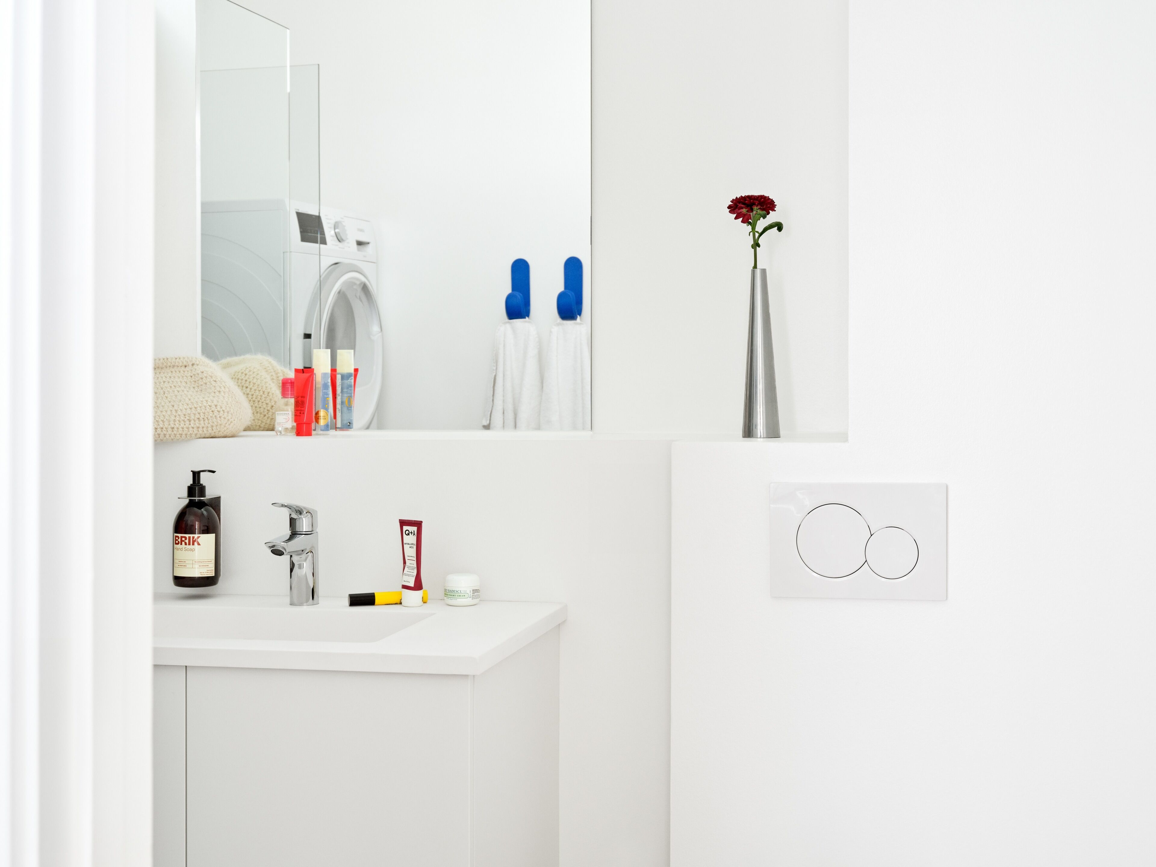 A modern bathroom with a white sink, a mirror, a washing machine, toiletries, and a vase with a flower.
