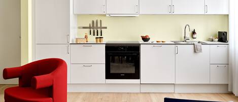 Una cocina moderna con armarios blancos, un horno negro y un lavamanos. Hay cuchillos colgados en la pared, una cafetera y un bol de fruta sobre la encimera.