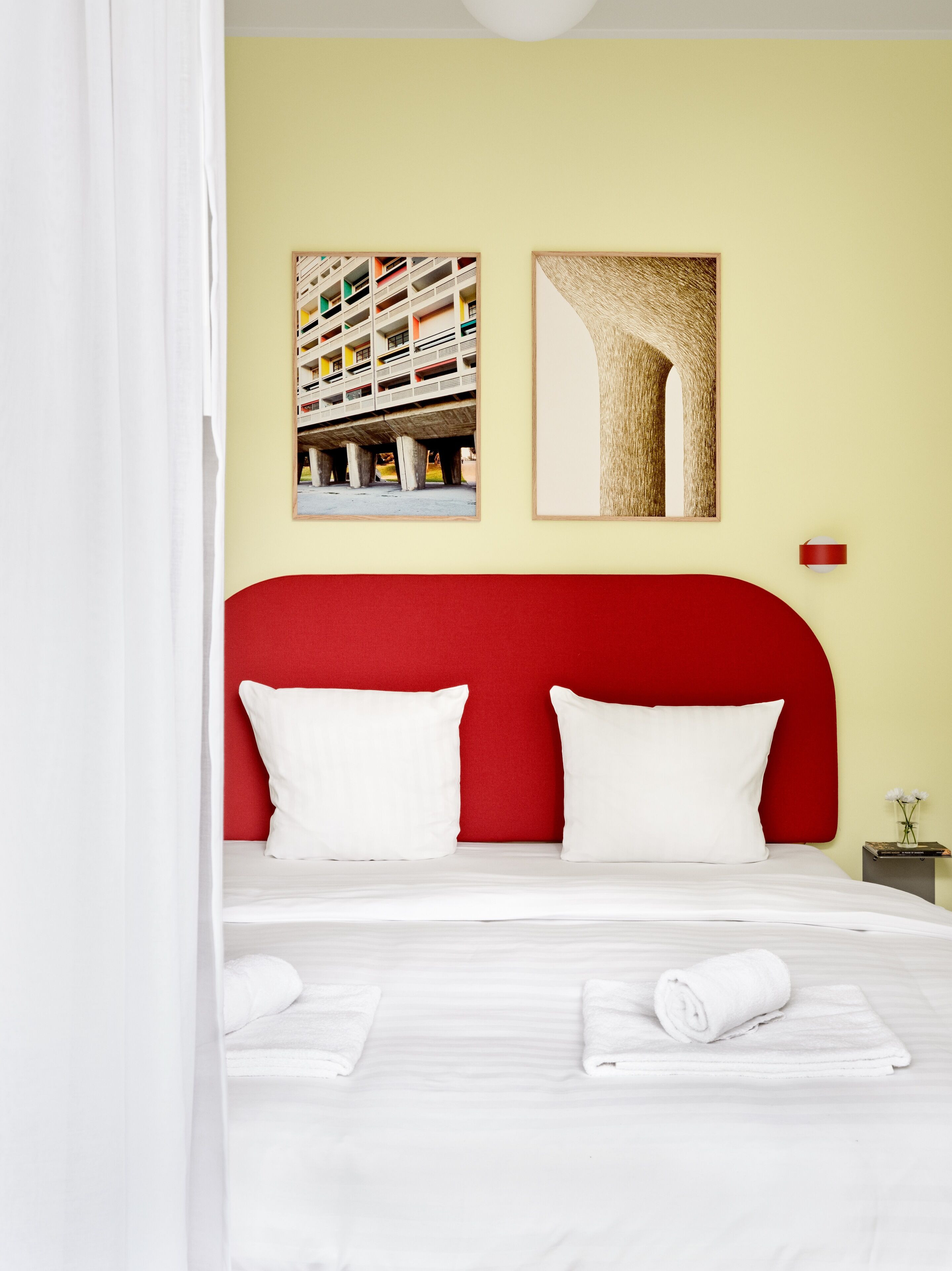 A hotel room with a red headboard, two white pillows, a neatly made bed with white linens, and two rolled towels.