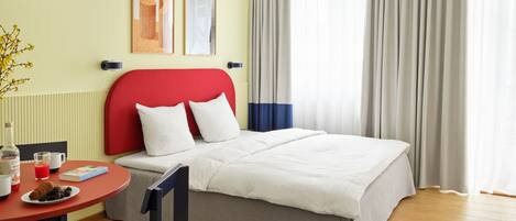 Habitación de hotel con una cama, una mesa redonda con cosas para el desayuno, una silla y una ventana con cortinas.