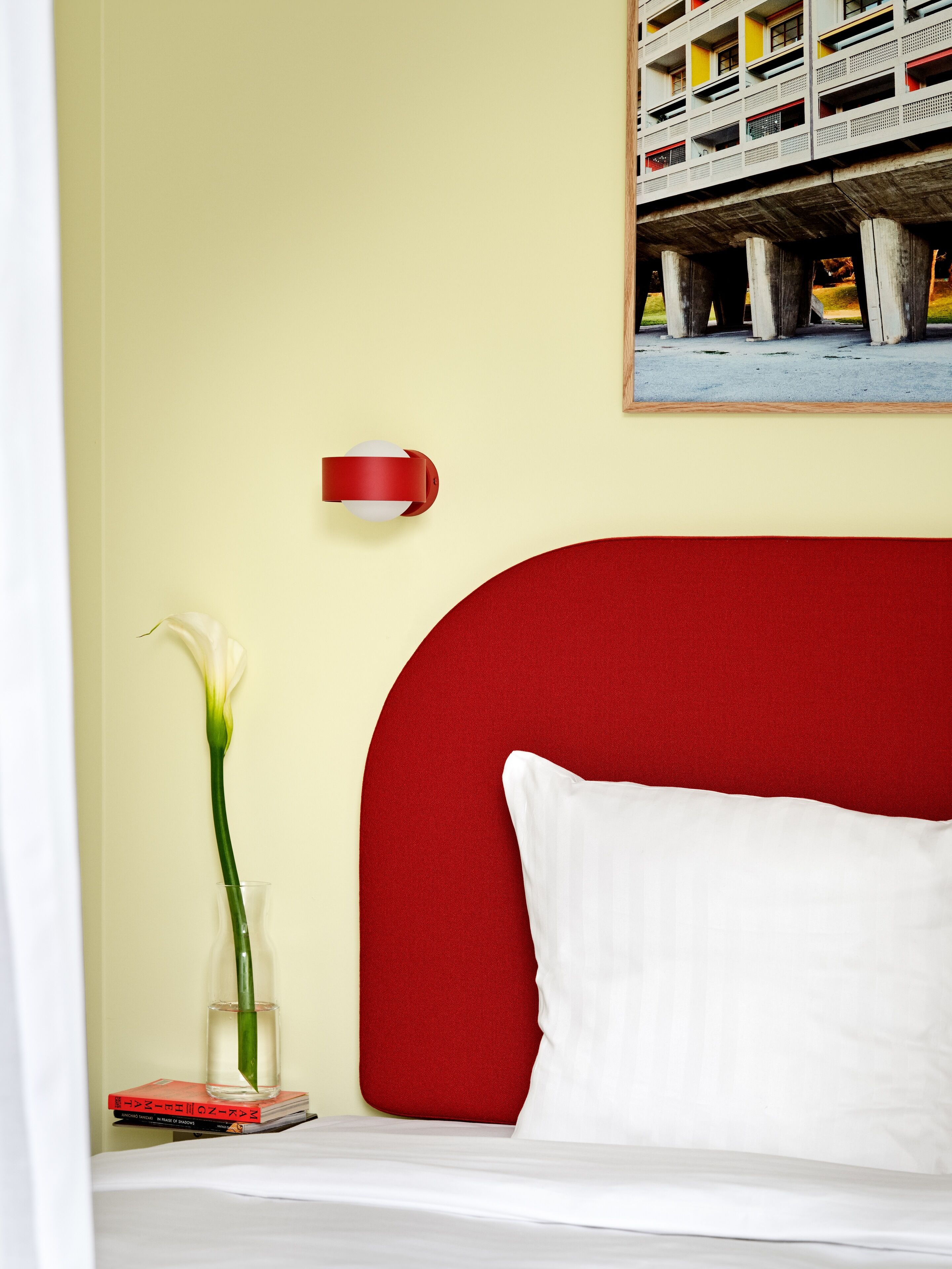 Un lit avec une tête de lit rouge, un oreiller blanc, un vase avec une fleur blanche et un tableau encadré accroché au mur.