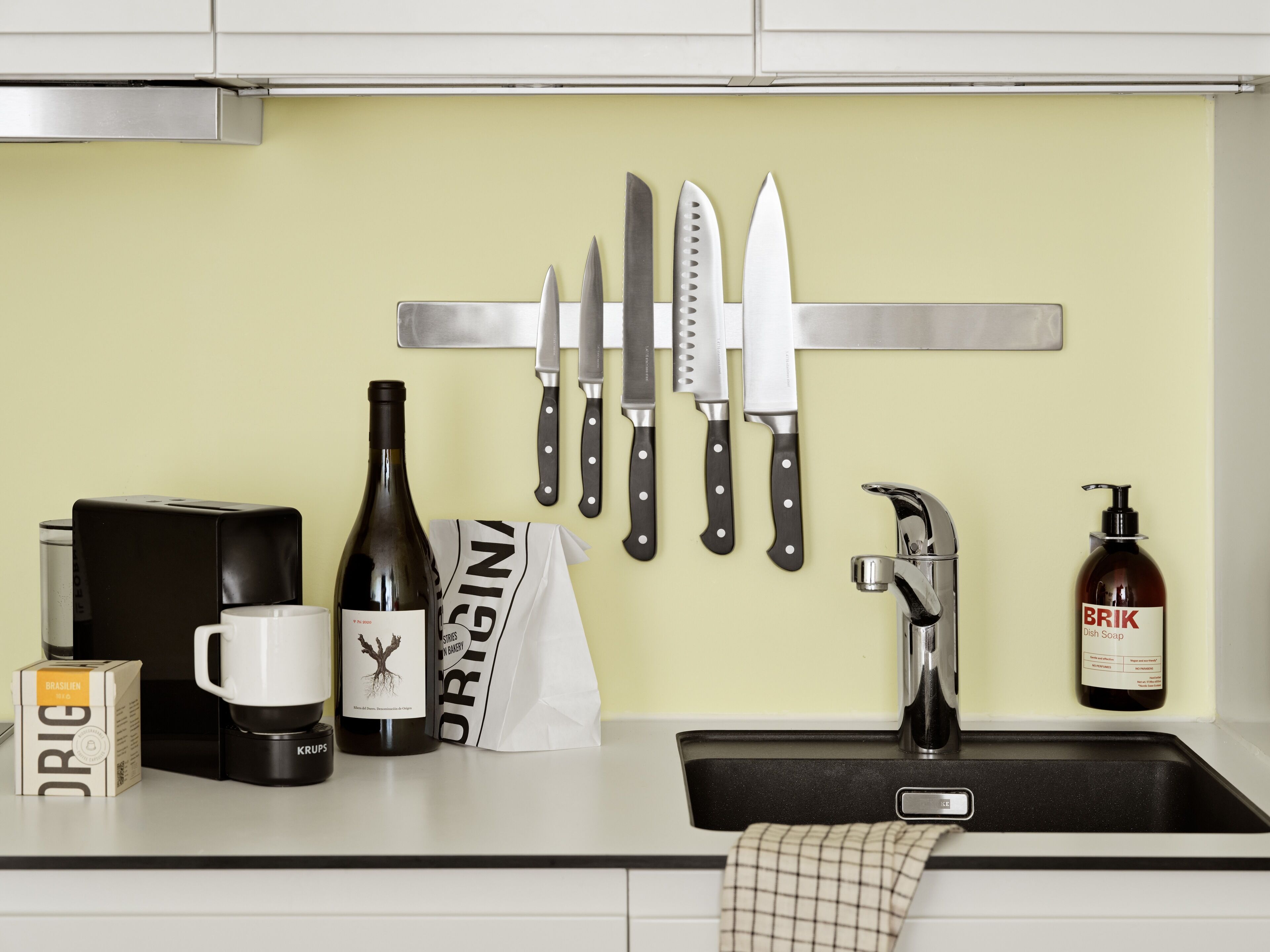 A kitchen counter with a coffee maker, a wine bottle, a bag of Origin brand coffee, a BRIK dish soap bottle, and a knife rack with various knives.
