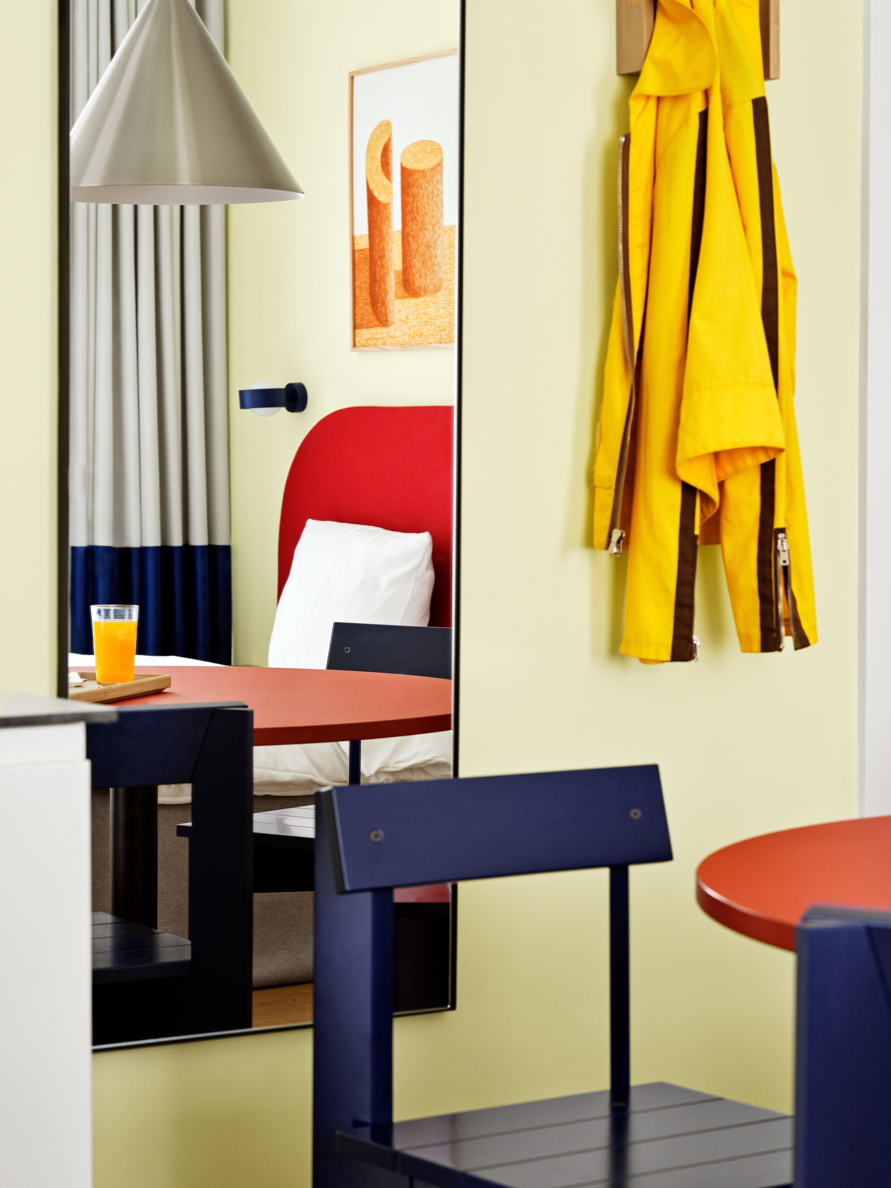 A hotel room with a bed, a table with a glass of orange juice, a chair, a lamp, and a coat rack with a yellow coat.