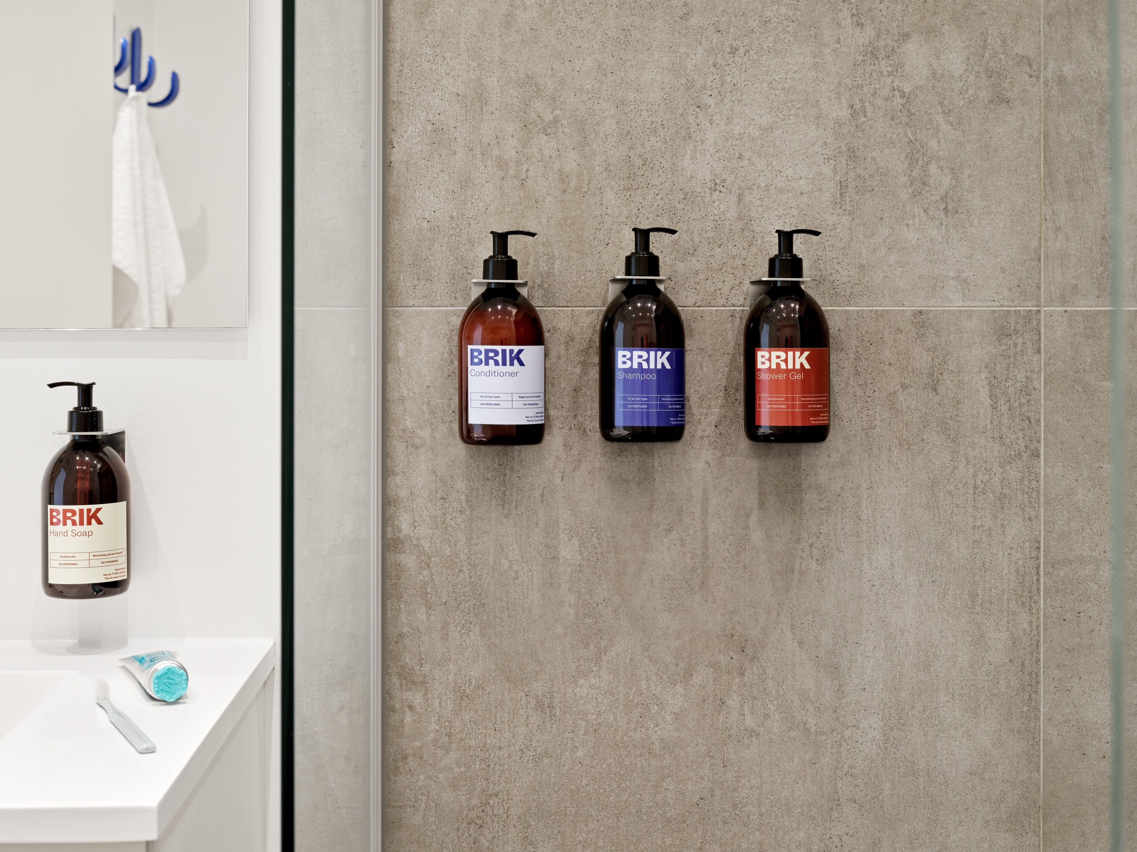 Een badkamer met een witte wasbak, een tandenborstel en vier BRIK toilettasflessen (handzeep, conditioner, shampoo en douchegel) aan de muur bevestigd.