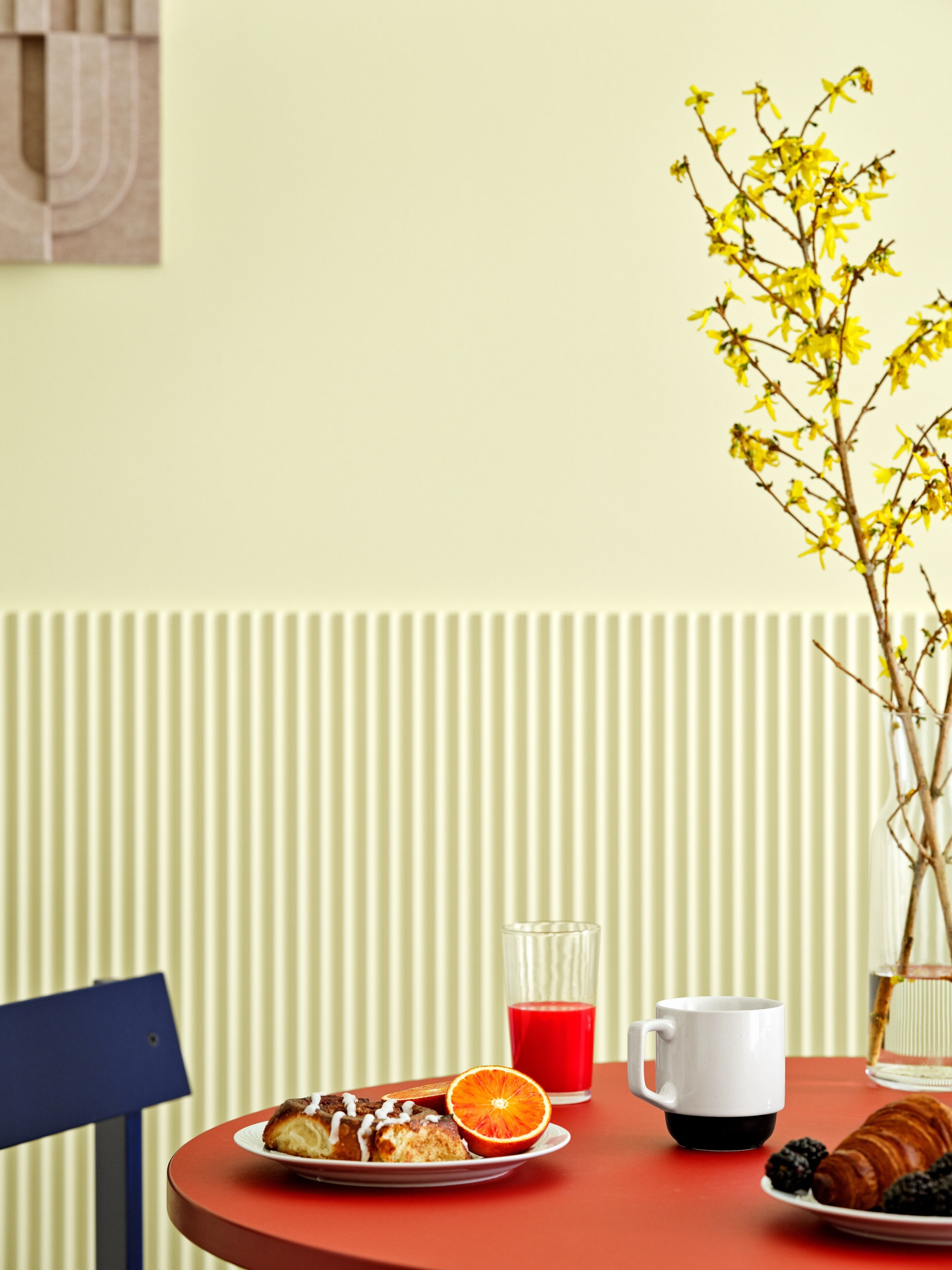 Ein Tisch mit Frühstücksangeboten, darunter ein Tablett Gebäck, ein Glas roter Saft, eine weiße Tasse und eine Vase mit gelben Blumen.