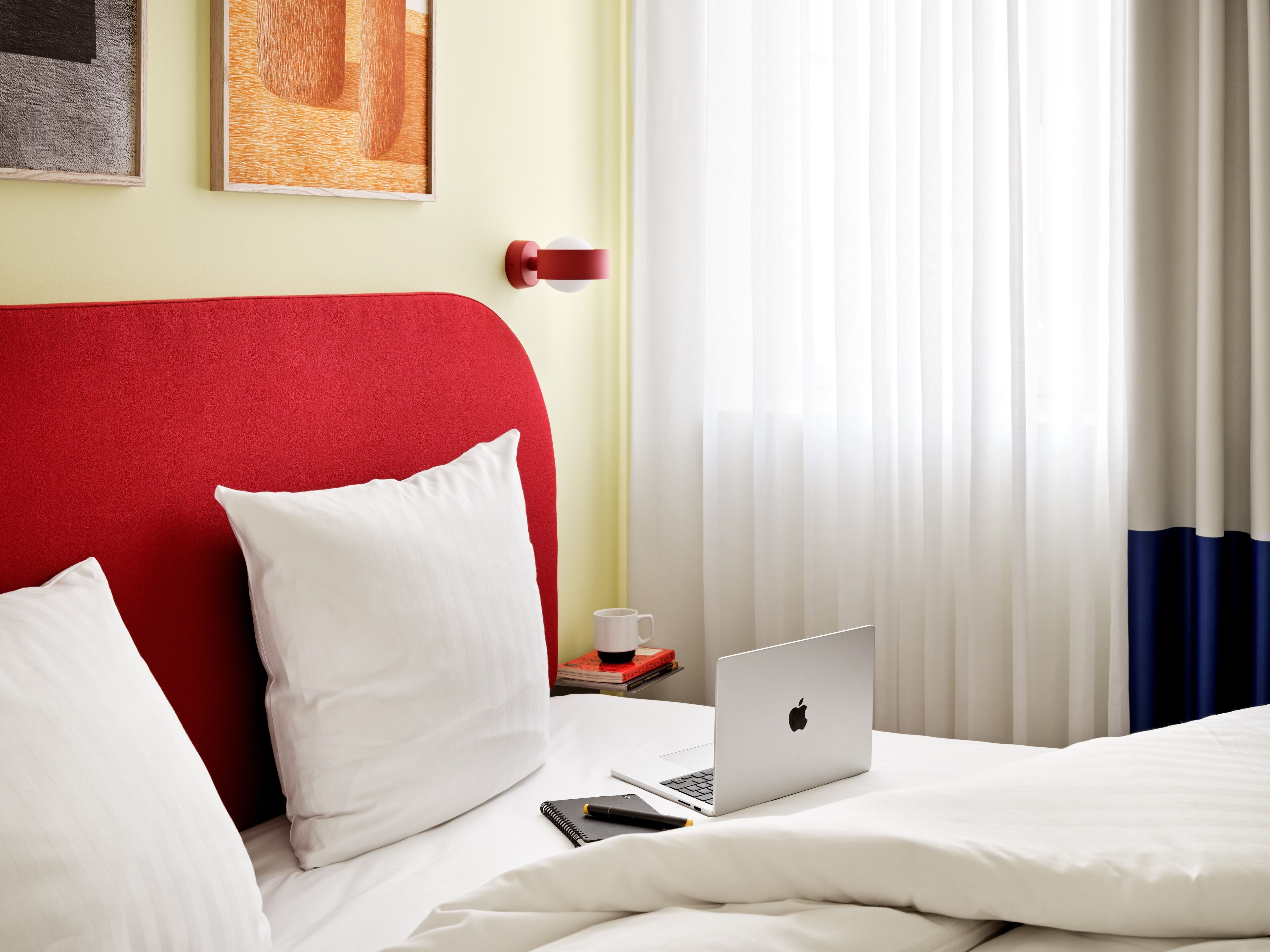 A hotel room with a red headboard, white bedding, a laptop on the bed, and a bedside table with a cup and a book.
