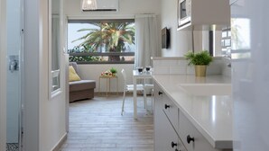 Een moderne keuken met witte kasten, een eethoek met een witte tafel en stoelen, en uitzicht op palmbomen door een raam.