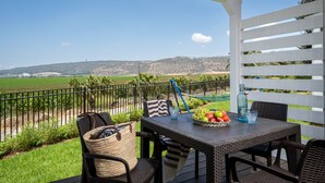 Een terras buiten met rieten meubels, een tafel met fruit en drankjes, en uitzicht op een groen veld en heuvels.