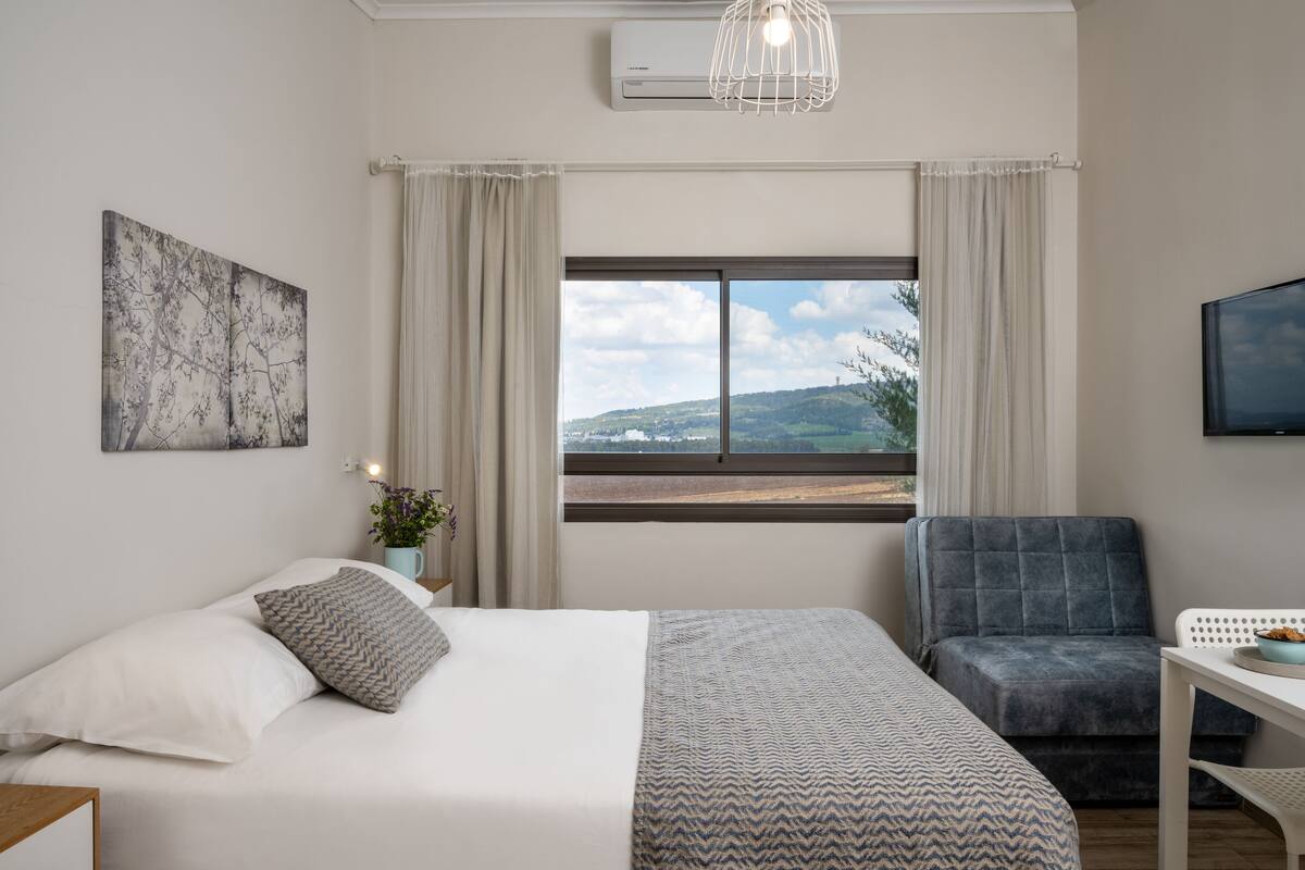 Ein Schlafzimmer mit Bett, Sessel, Tisch, Fernseher, Fenster mit Blick und Klimaanlage.