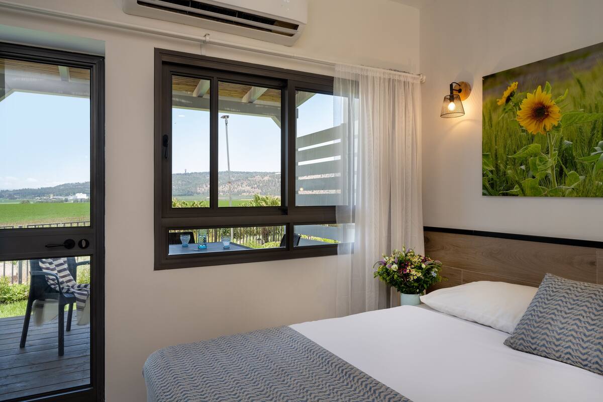 Ein Schlafzimmer mit einem großen Bett, einem Fenster mit Blick auf eine grüne Landschaft, einer Wandleuchte und einem Gemälde von Sonnenblumen.