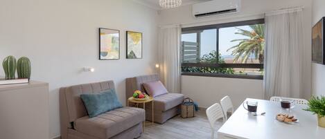 Una sala moderna con un sofá, una mesita con frutas, una mesa de comedor con sillas y un ventanal con vistas a palmeras.
