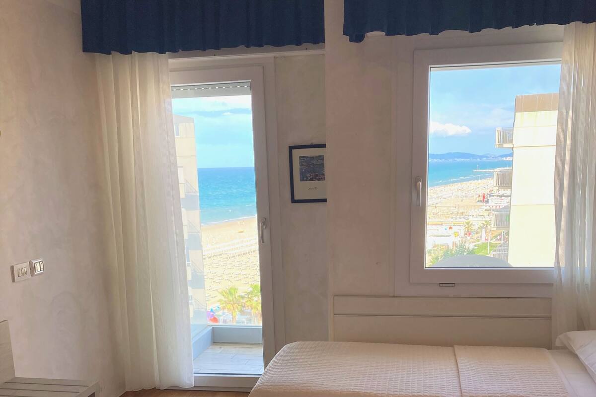 A bedroom with two beds, a window with a view of the beach, and a door leading outside.