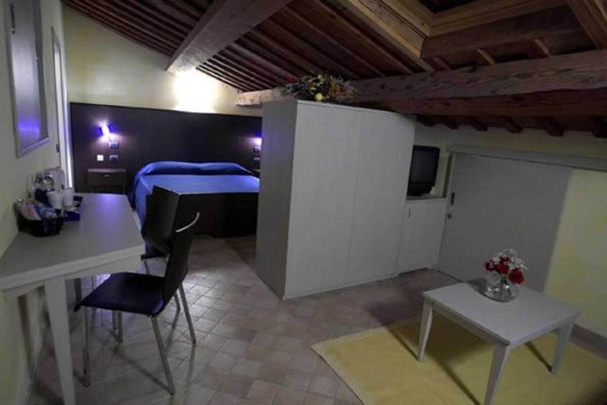 A compact room with a bed, a desk, a chair, a small table with a vase, and a television.