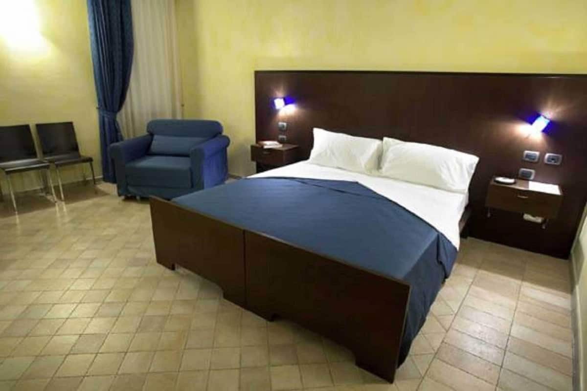 A hotel room with a large bed, a blue armchair, two bedside tables with lamps, and a window with curtains.