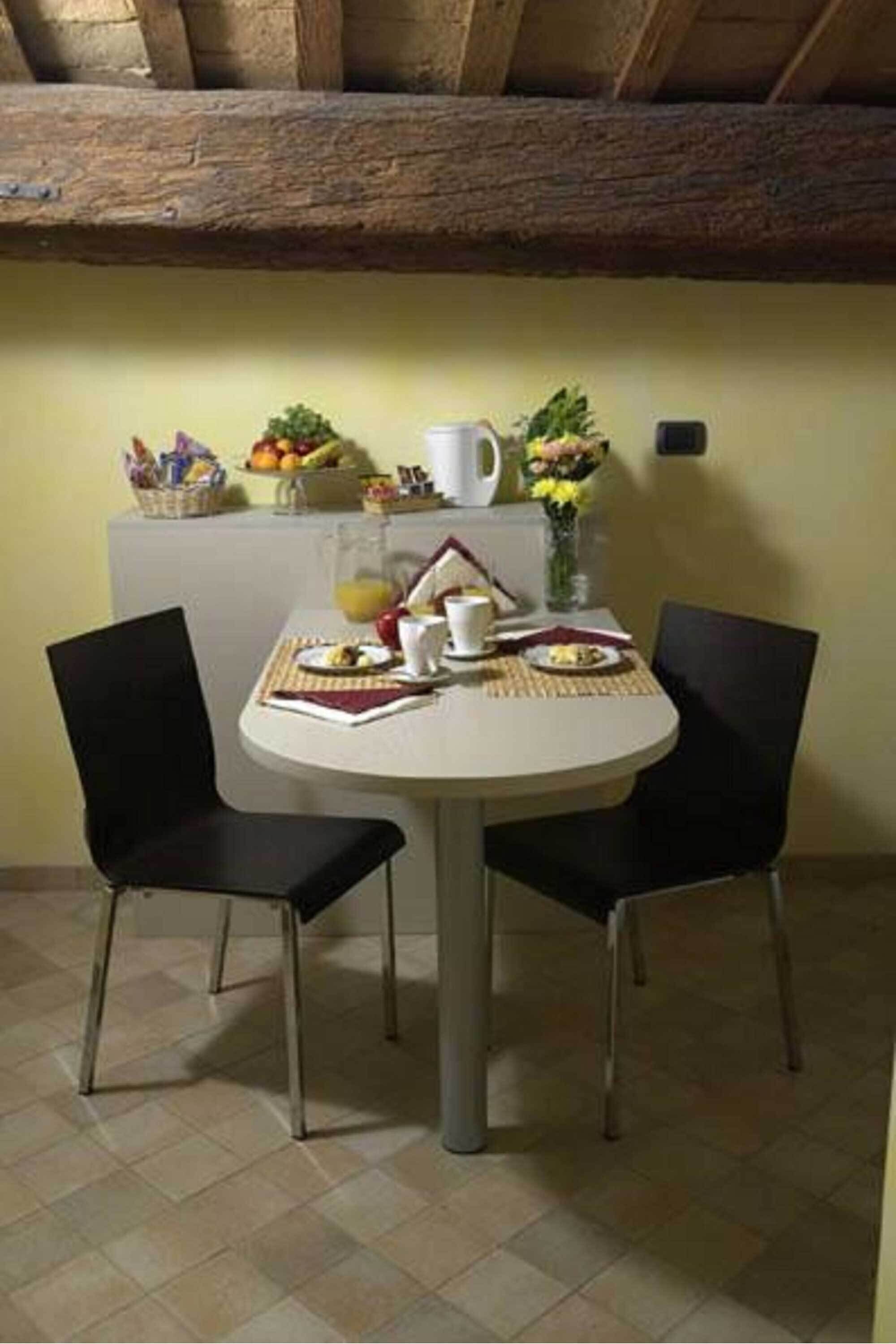 Comedor con mesa redonda, dos sillas negras y una disposición para el desayuno que incluye frutas, una taza de café y un jarrón con flores.