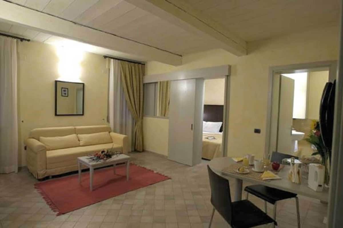 A compact living room with a sofa, a dining table, and a small coffee table. There is a bed in the adjacent room.