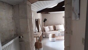 Ein schmaler Flur mit freiliegenden Steinwänden, einem weißen Heizkörper und einer Holzbalkendecke. Im Hintergrund ist eine gemütliche Wohnbereits mit einer Couch, Kissen und einem kleinen Tisch zu sehen.