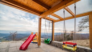 Una terraza de madera con un columpio y un tobogán rojo, con vistas a un paisaje montañoso.