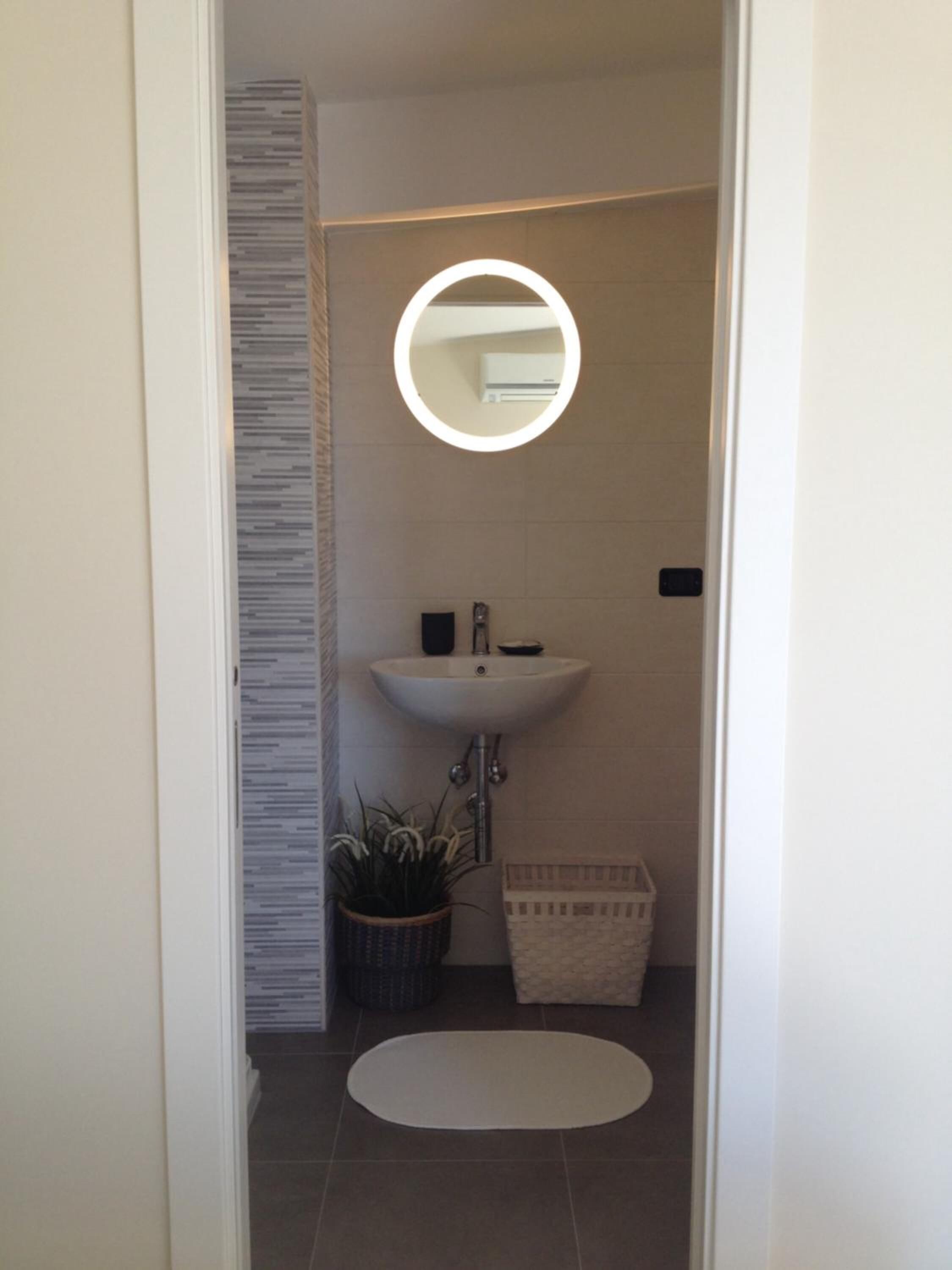 A narrow bathroom with a white sink, a round mirror, a potted plant, and a small storage basket.