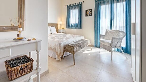Een slaapkamer met een groot bed, een nachtkastje, een rieten stoel en een raam met blauwe gordijnen.