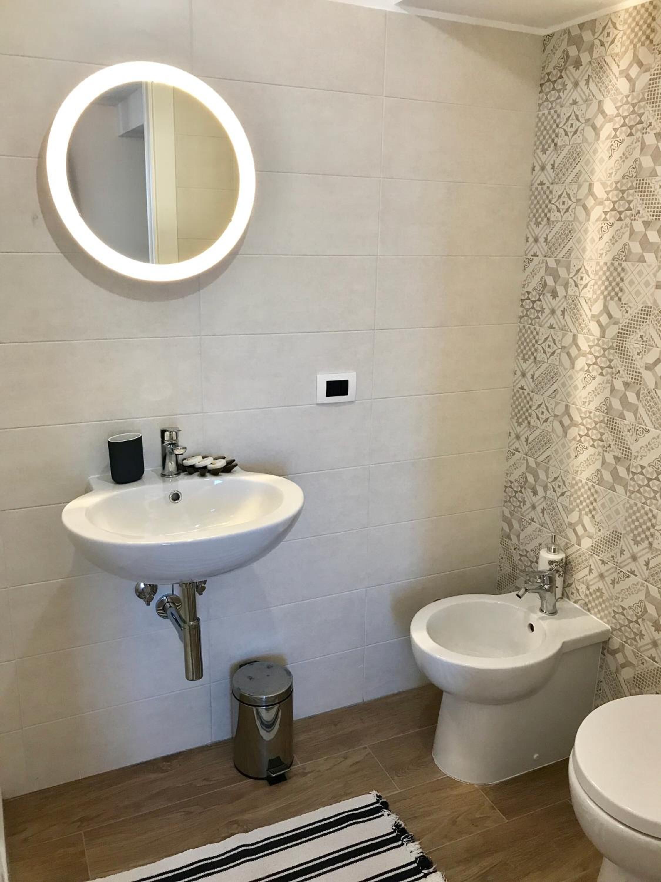 Et badeværelse med en hvid vask, en brusertoilet og et toilet. Der er en lille skraldespand og en stribet måtte på gulvet.