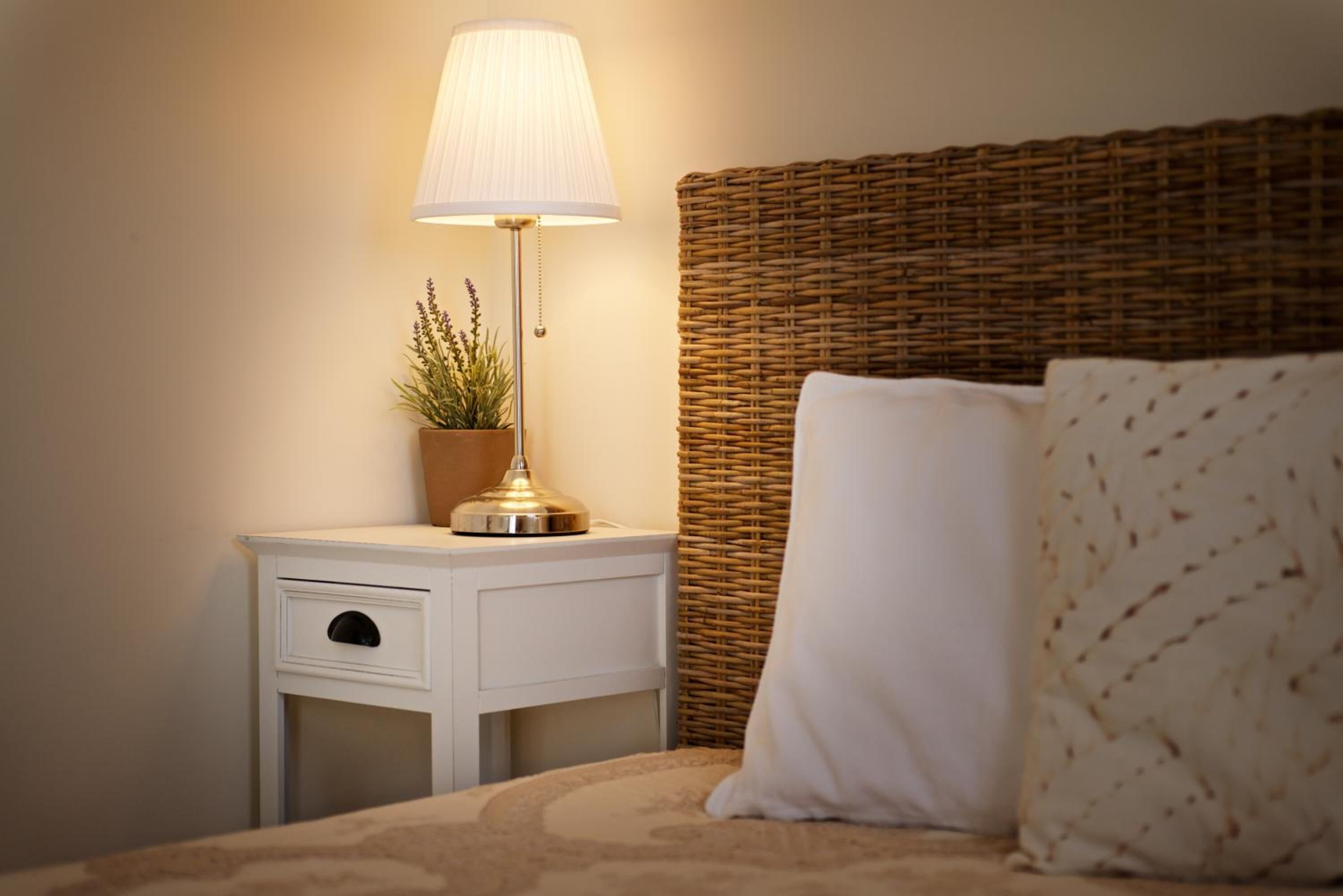 A neatly arranged bedroom with a bed, a bedside table with a lamp, a potted plant, and a wicker headboard.