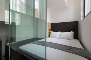 A modern hotel room with a glass partition, a large bed with white bedding, and a dark headboard.