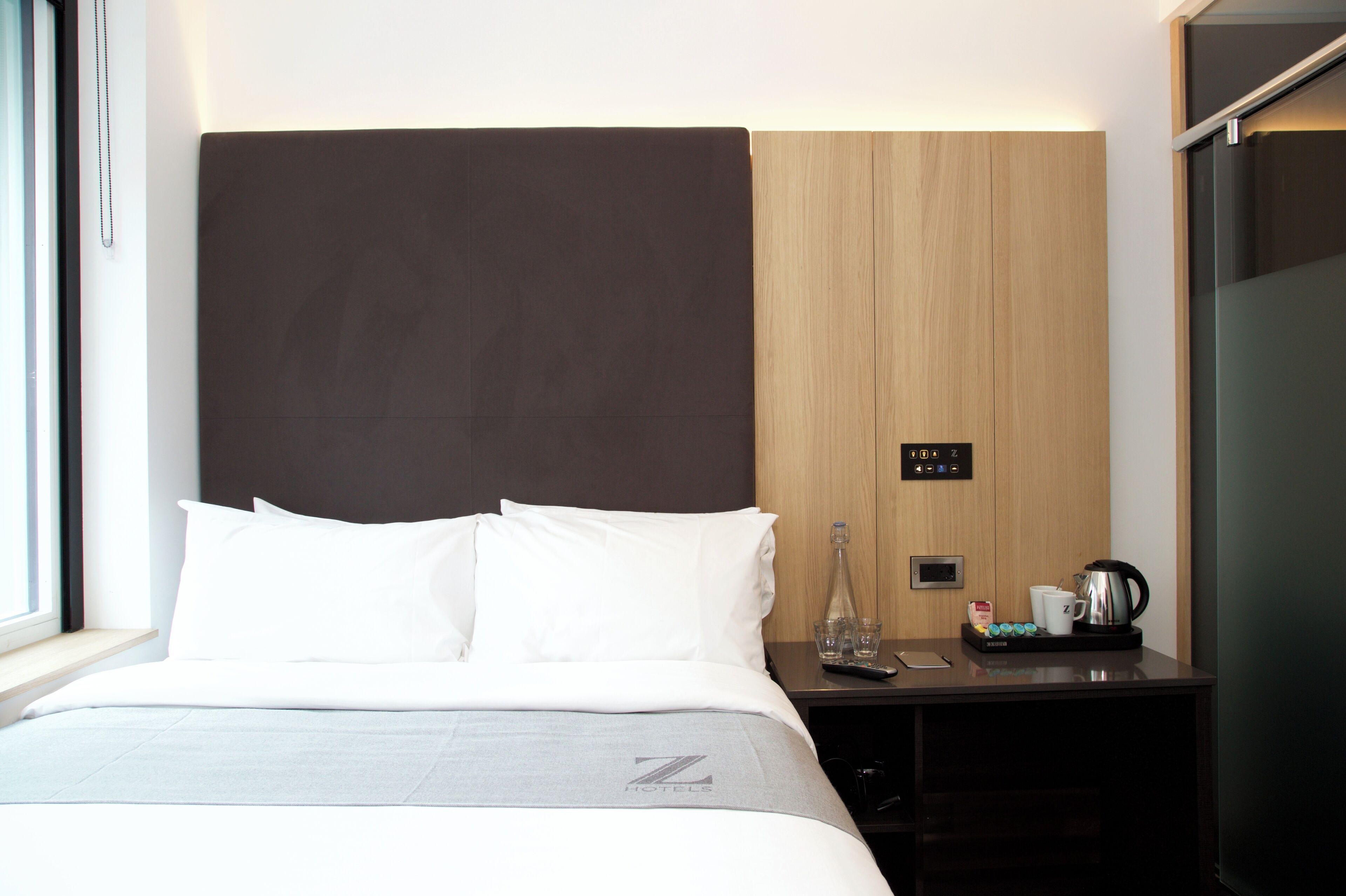 A modern hotel room with a large bed, a nightstand with a coffee maker, and a window with a view.