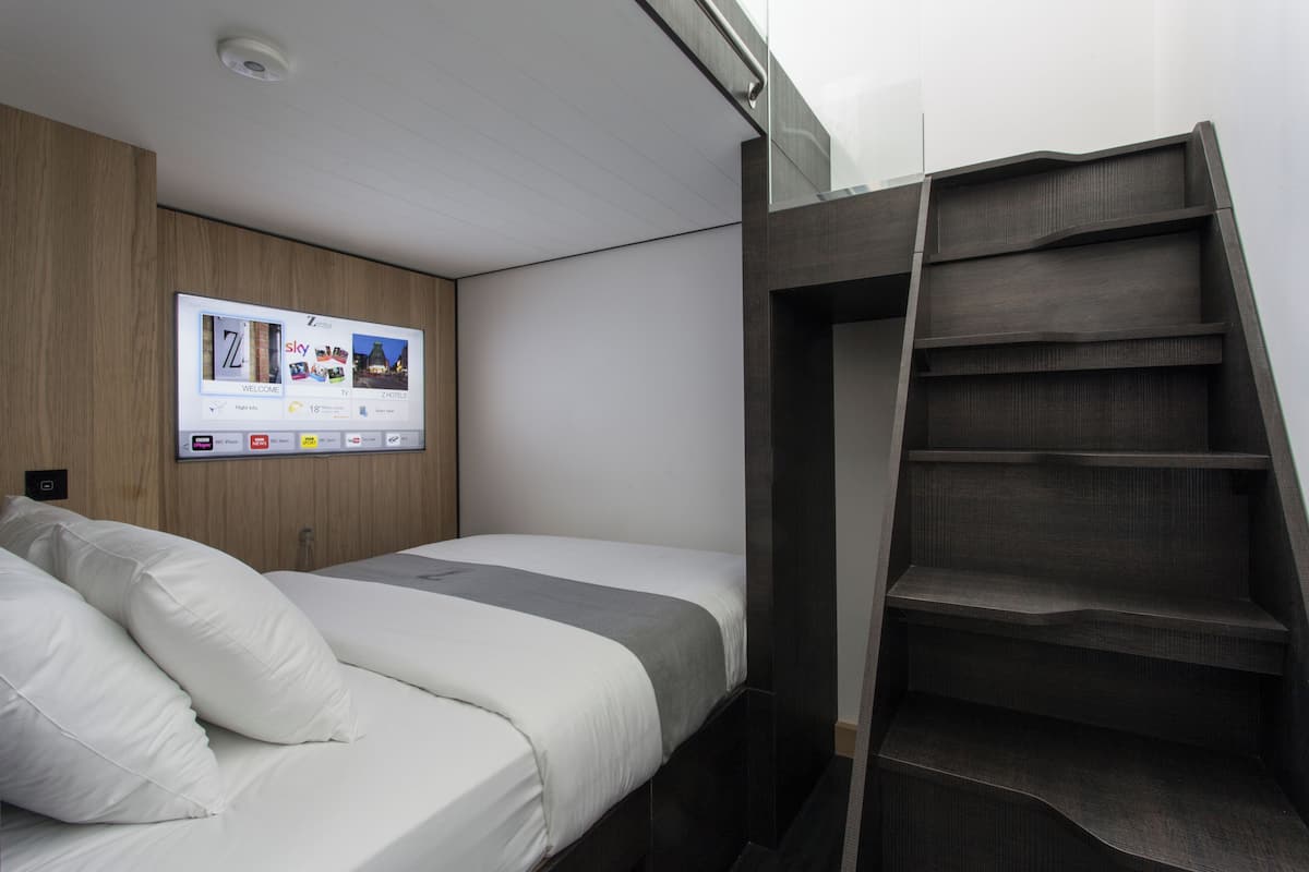 A modern hotel room with a bed, a digital display wall, and a built-in staircase.