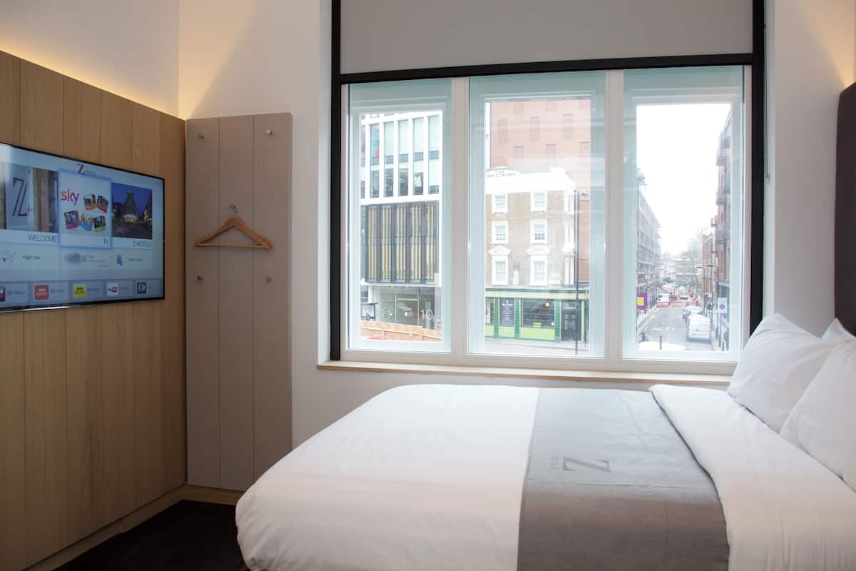 A hotel room with a large window, a bed, a TV mounted on the wall, and a wooden wardrobe.