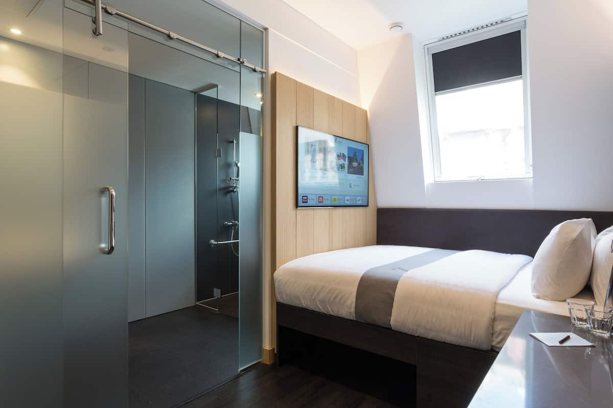 A modern hotel room with a bed, a TV mounted on the wall, a glass shower enclosure, and a window with a view.