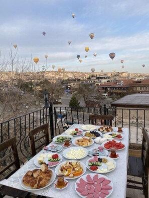 Une table dressée à l’extérieur, avec différents plats, donnant sur une ville où l’on aperçoit des bâtiments et des montgolfières dans le ciel.