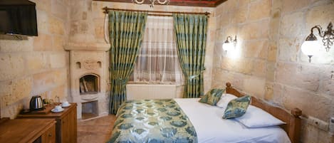 A bedroom with a bed, wooden headboard, a nightstand, a bedside table, a fireplace, and stone walls.